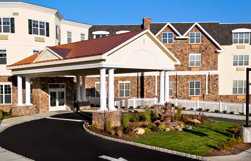 Front exterior view of The Bristal Assisted Living at Woodcliff Lake featuring a covered entrance with white columns, stone and beige facade, landscaped garden with a small fountain, and a driveway.