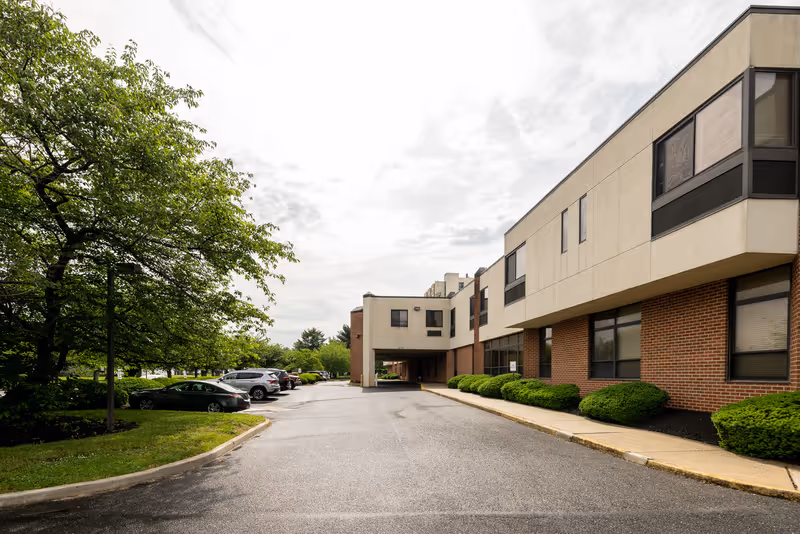 Exterior view of a two-story senior living facility building with a brick and beige facade, surrounded by neatly trimmed bushes and trees. Several parked cars are visible along the driveway leading to the entrance under a covered walkway. The sky is partly cloudy.