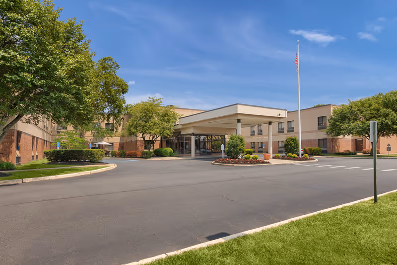 Entrance and driveway of a low-rise care facility with a covered porte-cochère, flagpole, and landscaped grounds.