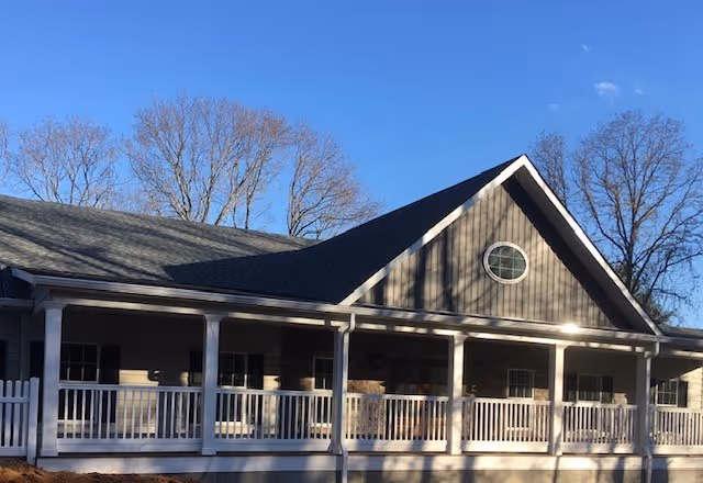 Front exterior of a single-story memory care building with a covered porch, white railing, gabled roof, and bare trees against a clear blue sky.