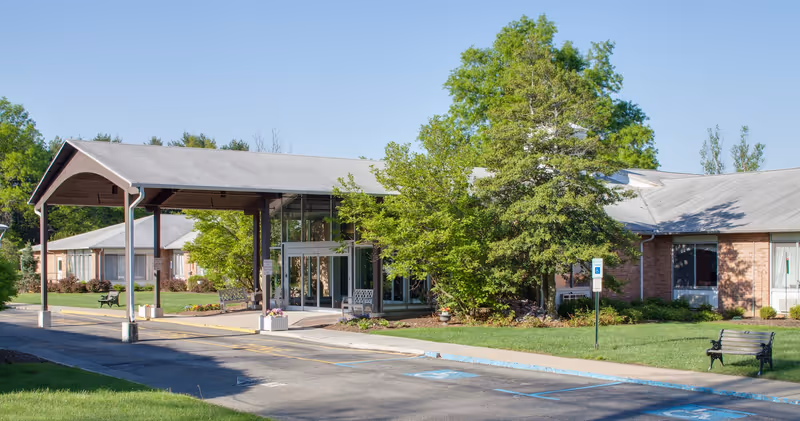 Exterior view of Holly Manor Center showing a single-story brick building with a covered entrance, surrounded by green trees and grass, with benches and a parking area including handicapped parking spaces.