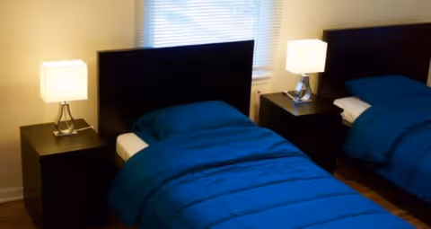 A bedroom with two single beds, each with a dark headboard and blue bedding. There are two matching bedside tables, each with a modern lamp. A window with blinds is visible behind the beds.