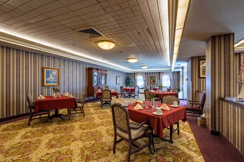A dining room with multiple tables covered in red tablecloths, each set with white napkins, cups, glasses, and silverware. The room features patterned carpet, striped wallpaper, framed paintings, and a wooden cabinet. Ceiling lights illuminate the space, and windows with curtains are visible in the background.