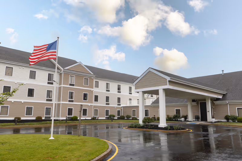 Exterior view of Mira Vie at Toms River assisted living facility showing a three-story building with beige and white siding, an American flag on a flagpole, a covered entrance, and a wet driveway under a partly cloudy sky.