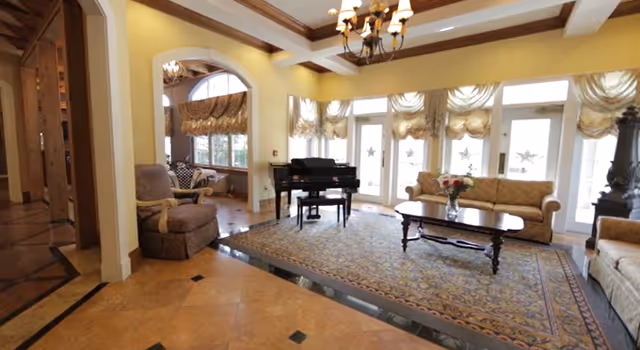 A spacious and elegant living room with a grand piano, a large patterned area rug, a wooden coffee table with a vase of flowers, and several upholstered sofas and armchairs. The room features large windows with decorative curtains allowing natural light to fill the space, and a chandelier hanging from the ceiling.