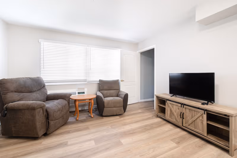 A bright living room with two brown recliner chairs separated by a small wooden side table, a large window with white blinds, a white door slightly open, and a wooden TV stand with a flat-screen television on top.