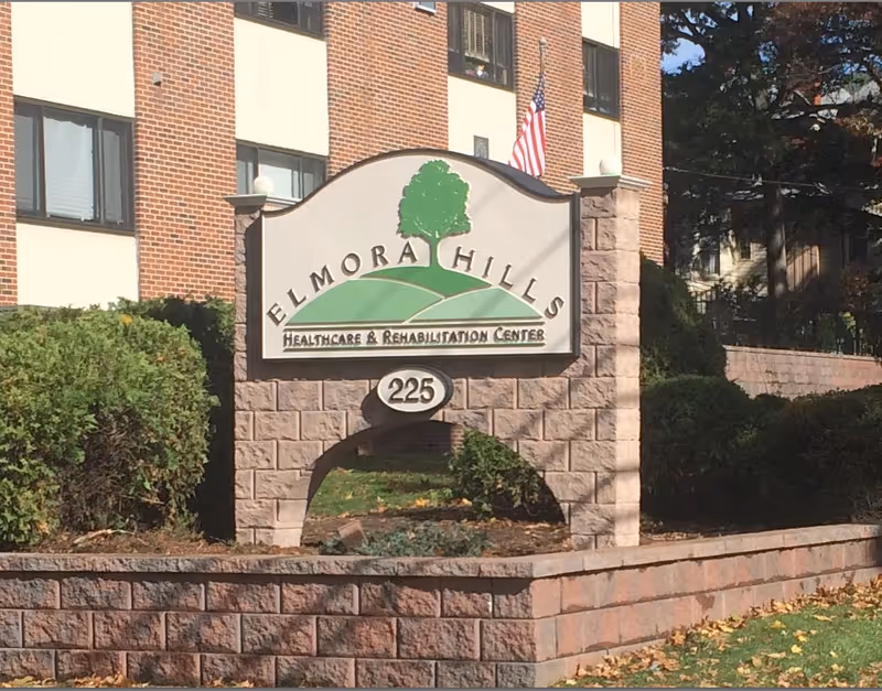 A stone sign in front of a brick building displaying the name Elmora Hills Healthcare & Rehabilitation Center with the number 225 below it. The sign features a green tree and hills graphic, with bushes and trees surrounding the area.