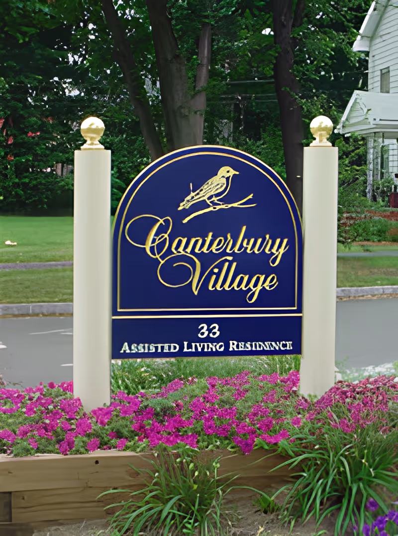 A blue and gold sign for Canterbury Village Assisted Living Residence, surrounded by purple flowers and greenery, with trees and a white building in the background.