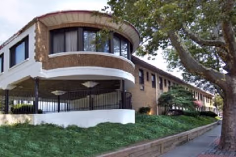 Exterior view of a brick multi-story building with a rounded corner, landscaped greenery and a tree-lined sidewalk.