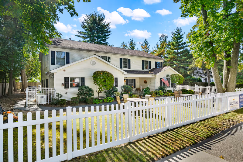 Exterior view of a two-story white building with black shutters surrounded by trees and a white picket fence. There is a small garden area with shrubs and a patio set with chairs in front of the building. An American flag is displayed near the entrance under a clear blue sky with some clouds.