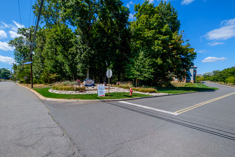 Street view of the entrance to Parker At Somerset facility with a landscaped area featuring a sign surrounded by trees and greenery under a blue sky with some clouds.