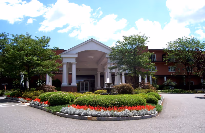 Front exterior view of Heath Village Retirement Community featuring a large entrance with white columns, a covered portico, a central fountain, well-maintained landscaping with green bushes and red flowers, and trees on either side under a partly cloudy blue sky.