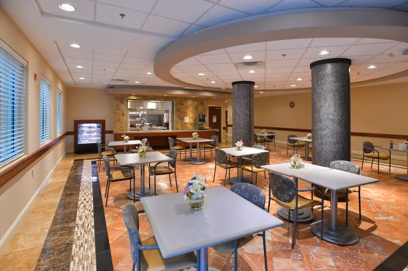 A dining room with several square tables and chairs arranged neatly. Each table has a small flower arrangement with an American flag. The room has large windows with blinds on one side, a tiled floor with a decorative border, and two large cylindrical columns. In the background, there is a serving window to the kitchen area and a refrigerated display case.