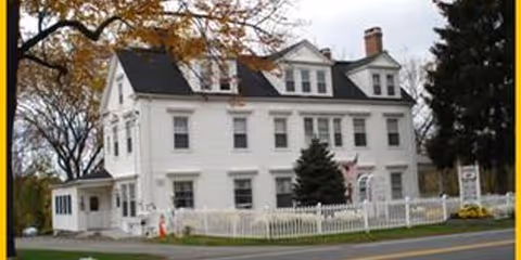 A large white multi-story house with a black roof, surrounded by a white picket fence and some trees, located next to a road. The building appears to be a senior care facility named Merry Heart Senior Care Services.
