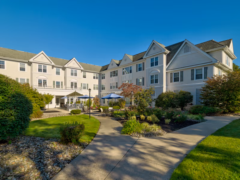Exterior view of The Residence at Cherry Hill, showing a large, multi-story building with white siding and multiple windows. In front of the building is a landscaped garden with bushes, trees, and a paved walkway leading to outdoor seating areas with tables and umbrellas under a clear blue sky.