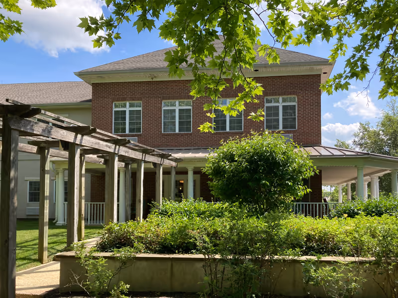 Exterior view of a two-story brick building with white columns and multiple windows, surrounded by green bushes and trees under a blue sky with some clouds. A wooden pergola structure is visible on the left side of the image.