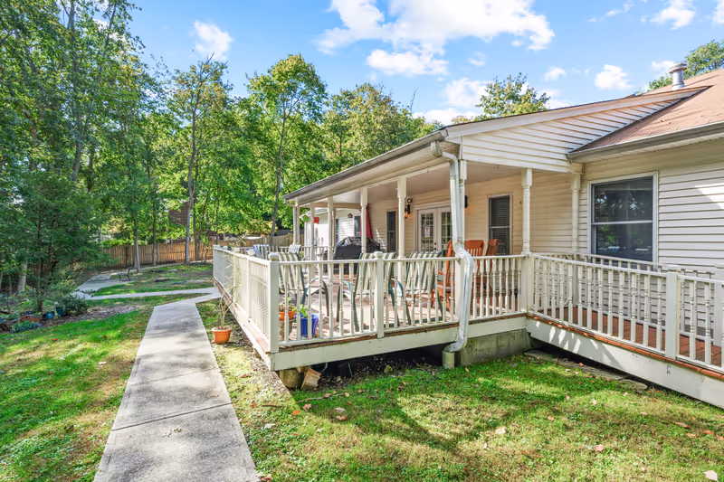 Single-story exterior of a memory care facility showing a covered porch with railings and chairs, a concrete walkway, and a grassy yard with trees.
