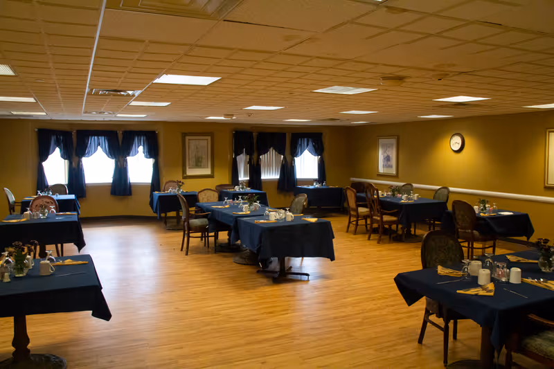 A dining room with several tables covered in dark blue tablecloths, each set with cups, utensils, and small flower arrangements. The room has wooden flooring, yellow walls, framed artwork, a clock, and windows with dark blue curtains.