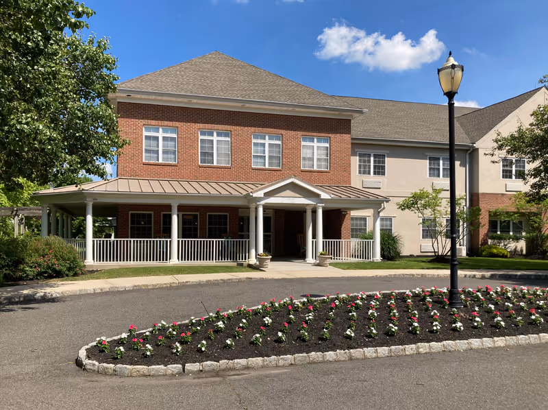 Exterior view of a two-story senior living facility building with a brick and beige facade, a covered entrance supported by white columns, a flower bed with pink and white flowers in front, a black street lamp, and trees on either side under a blue sky with a few clouds.