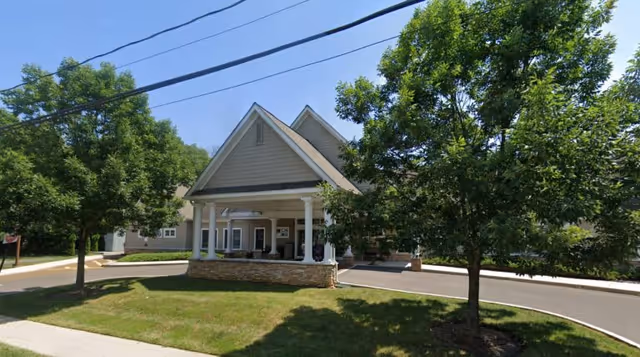 Front exterior view of Abrams Residence, a building with a covered entrance supported by white columns, surrounded by green trees and a well-maintained lawn under a clear blue sky.