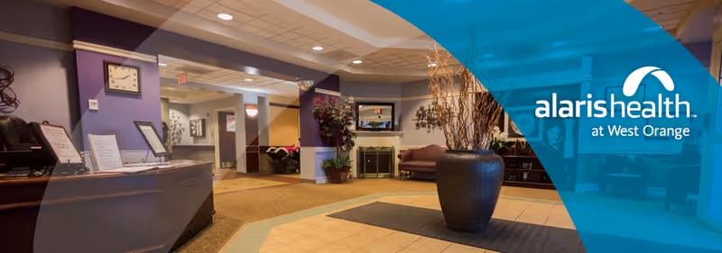 Reception area of Alaris Health at West Orange featuring a front desk with computer monitors, a clock on the wall, indoor plants, a large decorative vase with branches, seating area with couches, and warm lighting.