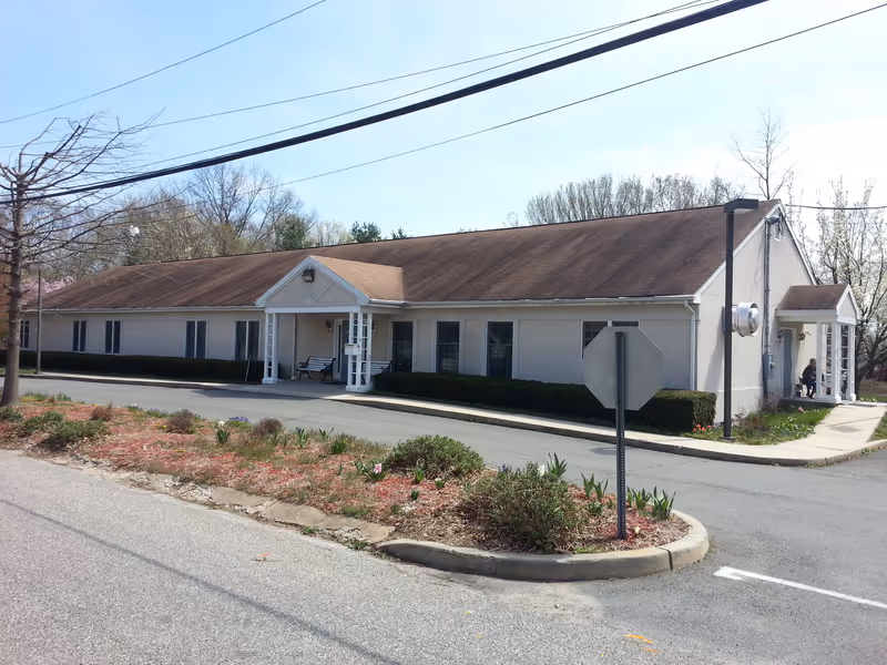 Exterior view of a single-story building with a brown roof and beige walls, featuring two small covered entrances with white pillars. There are benches near the entrances and a stop sign in the foreground. The building is surrounded by a paved driveway and some landscaping with bushes and small plants. Trees and a clear sky are visible in the background.