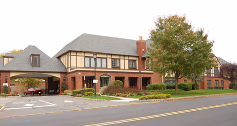 Exterior view of a large brick and timber senior living facility building with a covered driveway entrance, landscaped garden with bushes and a tree, and a paved road in front.