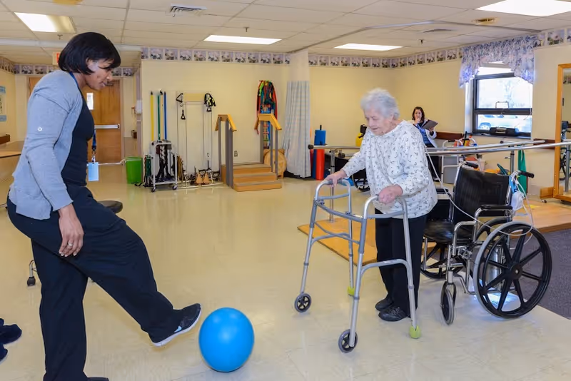 An elderly woman using a walker is playing with a blue ball indoors, assisted by a caregiver who is kicking the ball towards her. A wheelchair is nearby, and another person is seated in the background. The room appears to be a rehabilitation or therapy area with exercise equipment and handrails.