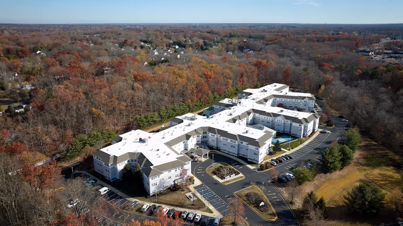 Aerial view of Monarch Bella Terra, a large senior living facility surrounded by trees with autumn foliage. The building has a white roof and multiple wings with parking lots and driveways around it.