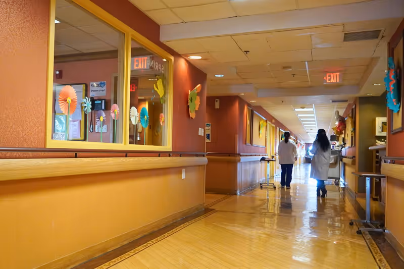 A brightly lit hallway in a senior care facility with polished wooden floors and warm-colored walls decorated with colorful paper flowers. Two staff members in white coats walk down the corridor, which has handrails on both sides and exit signs visible on the ceiling.
