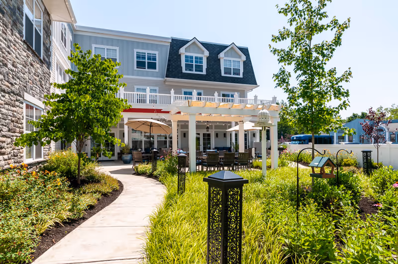 Outdoor garden area at Arbor Terrace Mountainside featuring a paved walkway, green plants, trees, decorative black lanterns, a white pergola with string lights, patio tables with umbrellas, and a multi-story building in the background under a clear blue sky.