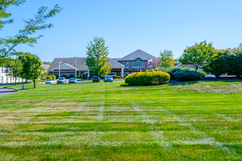 Wide view of a senior living facility building with a well-maintained green lawn in the foreground, several trees and bushes, and a few parked cars near the entrance under a clear blue sky.