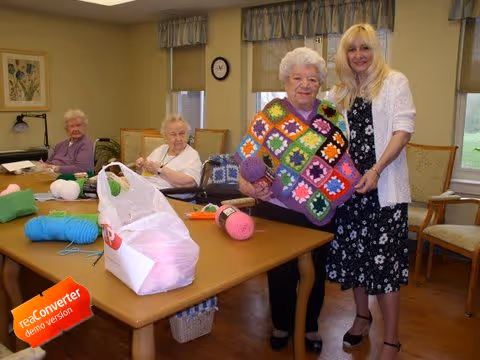 Three elderly women sitting around a table with yarn and knitting supplies in a well-lit room, while a younger woman stands next to one of the elderly women who is holding a colorful crocheted blanket.