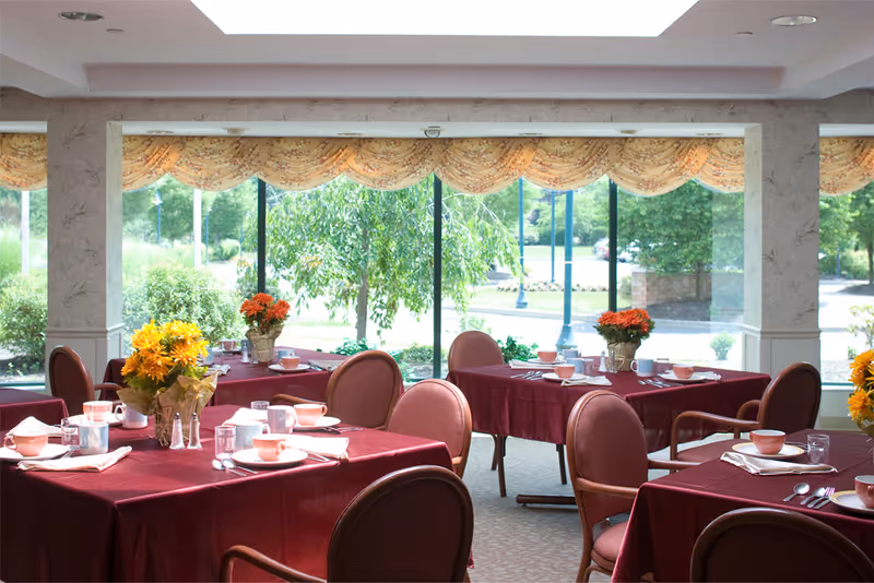 Dining room with several tables covered in burgundy tablecloths, each set with cups, glasses, plates, and silverware. There are flower arrangements on the tables, and large windows in the background show greenery and trees outside.