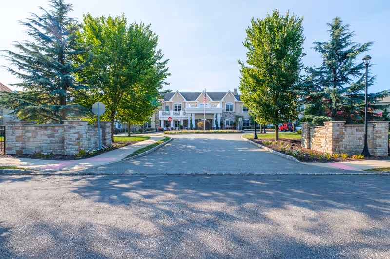 Front exterior view of Brandywine Livingston by Monarch facility with a driveway entrance flanked by stone walls and trees, leading to a two-story building with a covered entrance and an American flag.