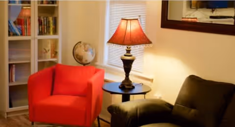 A cozy living room area with a red armchair, a black leather sofa, a small round side table with a decorative lamp, a globe on a shelf, and a window with blinds.