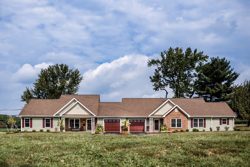 Single-story duplex-style building with two attached garages, front porches, and a large lawn under a cloudy sky.