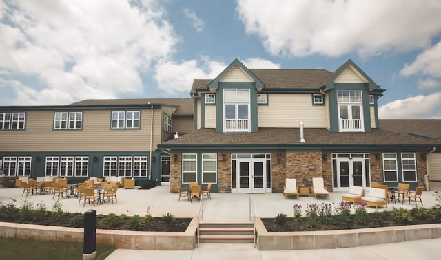 Front exterior of a two-story senior living building with a large patio, outdoor seating, and landscaped planters.