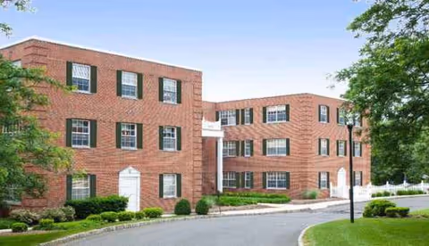 Exterior view of a three-story brick building with multiple windows, green shutters, and white doors, surrounded by greenery and a paved driveway under a clear blue sky.