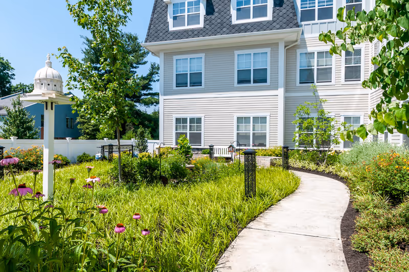 A sunny outdoor garden area at Arbor Terrace Mountainside with a curved concrete pathway surrounded by green plants and flowers. There is a white birdhouse on a post, a white bench near the building, and a beige multi-story building with multiple windows in the background.