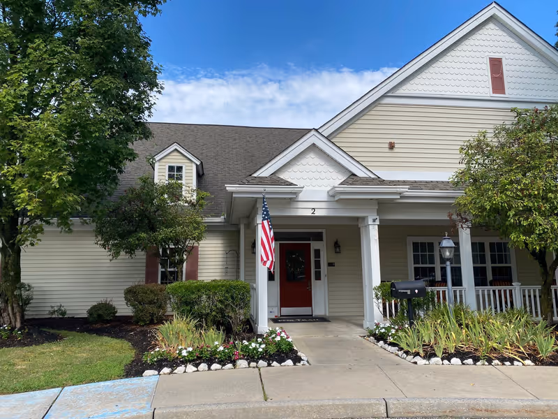 Exterior view of a single-story senior living facility building with beige siding, a red front door, white columns supporting a small porch, an American flag hanging near the entrance, landscaped flower beds, and trees on either side under a partly cloudy blue sky.