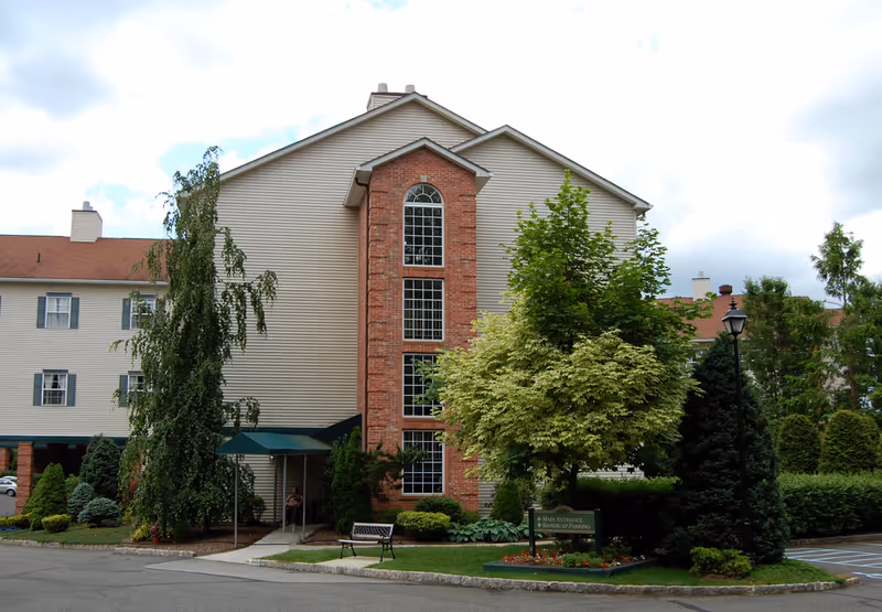 Exterior view of a multi-story assisted living facility building with beige siding and a central brick section featuring tall windows. The entrance has a green awning, and there are trees, shrubs, a bench, and a lamp post in the landscaped area near the entrance. A sign indicates the main entrance and handicap parking.