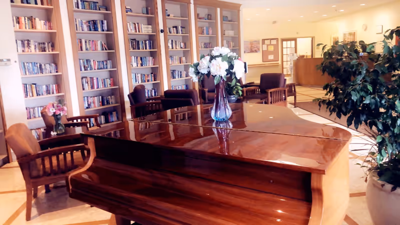 A polished wooden grand piano with a vase of white flowers on top is placed in a cozy room with bookshelves filled with books along the walls. Several wooden chairs with cushions are arranged around the room, and there is a large potted plant on the right side. The background shows a reception desk and a bulletin board on the wall.
