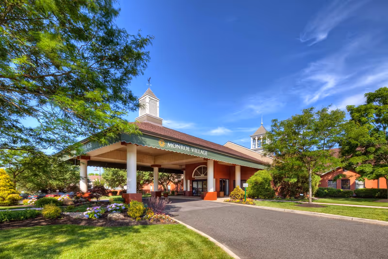 Front entrance of a senior living community with a covered porte-cochere, white columns, landscaped flower beds, and a blue sky.
