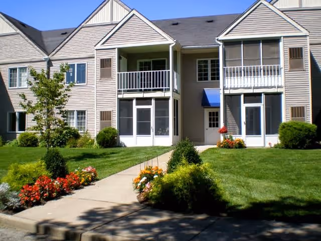 Front view of a two-story retirement community building with balconies, screened porches, a central walkway, and landscaped lawn with flower beds.