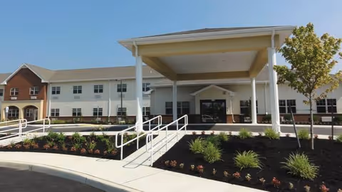 Front entrance of a two-story senior care building with a covered porte-cochère, accessibility ramps and landscaped beds under a clear blue sky.