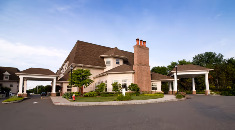 Exterior view of a senior living facility building with a large covered entrance and driveway. The building has beige siding, a brown roof, multiple windows, and a prominent brick chimney. There are landscaped areas with green bushes and trees around the building, and the sky is clear with some clouds.