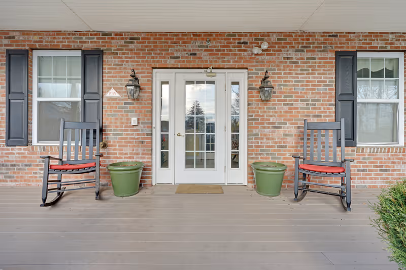 Front porch of a brick building with two black rocking chairs with red cushions, two green plant pots, and a white double door with glass panels. Two windows with black shutters flank the door, and two wall-mounted lantern-style lights are above the plant pots.