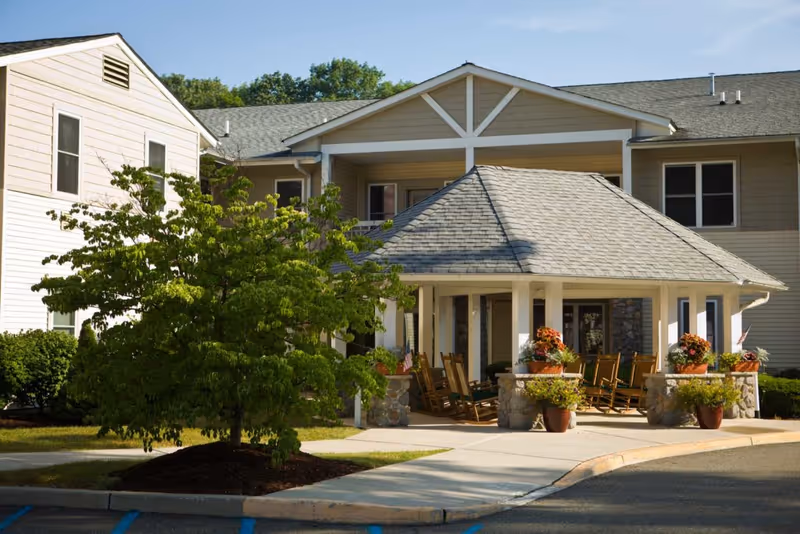 Exterior view of a senior living facility with a covered porch area featuring several wooden rocking chairs and potted plants. The building has beige siding, multiple windows, and a gray shingled roof. A tree and landscaped greenery are visible in the foreground under a clear sky.