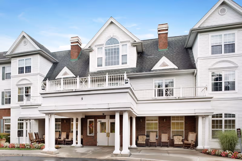 Front exterior view of a senior living facility building with white siding, brick accents, multiple windows, and a covered entrance supported by white columns. Several rocking chairs are placed under the covered area, and there are flower beds with colorful flowers along the front.
